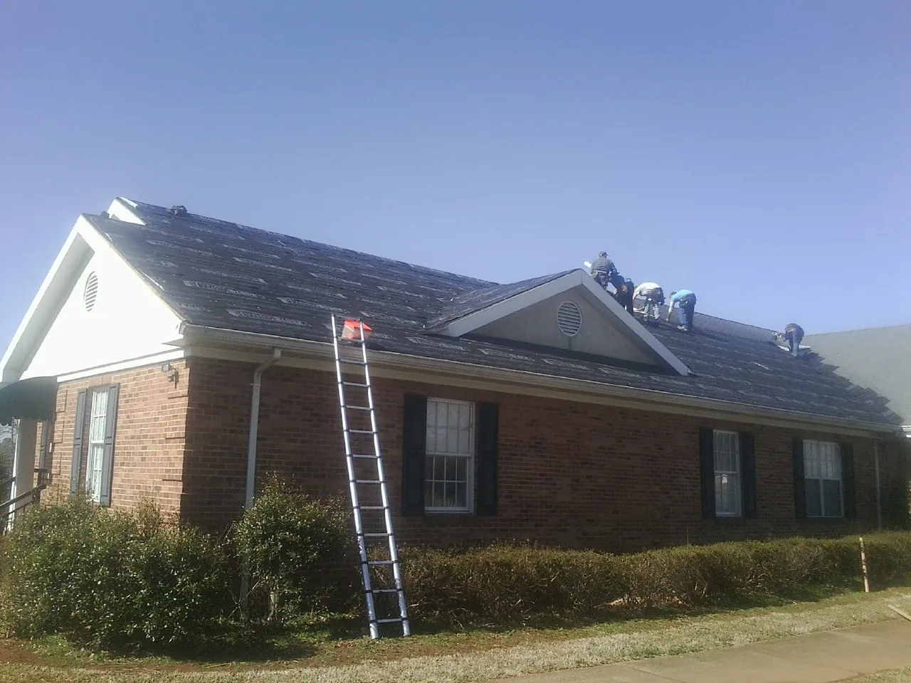 Crew tearing shingles off a brick ranch roof with a ladder set — Metal Roof Repair work by our crew in Poplar Grove