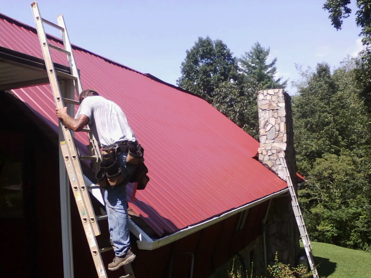 Roofer on a ladder beside a red metal roof and stone chimney — Soffit & Fascia Repair by Roofing Brothers in Poplar Grove